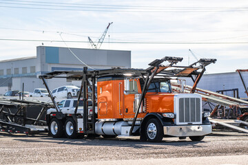 Extended cab orange big rig classic car hauler semi truck with semi trailer standing on the industrial parking lot