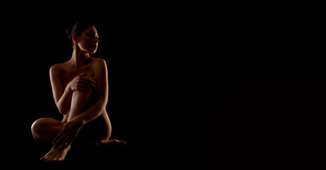 A woman sits alone in a dark room, her arms crossed and her legs crossed. The only light is from the side, illuminating her profile and creating a sense of mystery, copy space