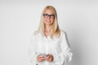 © Prostock-studio - A blonde woman in her 40s or 50s wearing glasses and a white shirt is holding a smartphone in her hands. She is looking directly at the camera and smiling. The background is a plain white wall.