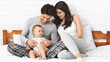 © Prostock-studio - A happy family sits on a white bed in a bedroom. The father is holding their baby, and the mother is smiling at her family. They are all wearing casual clothes, and they look very comfortable.