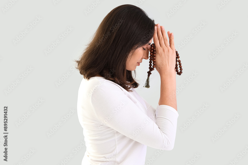 Young woman praying with beads on light background