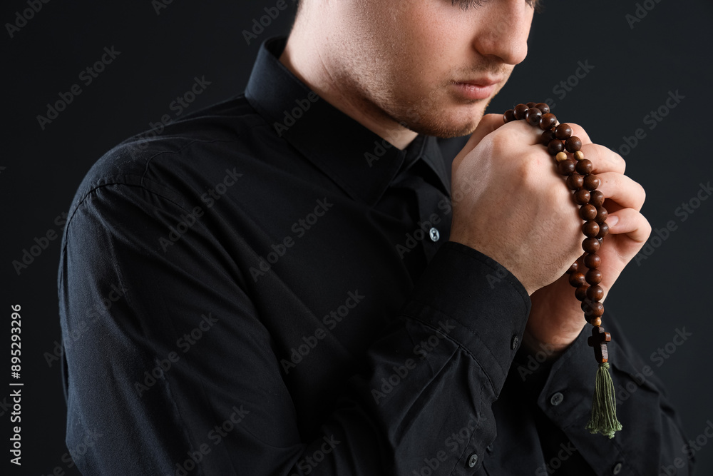 Young man praying with beads on dark background, closeup