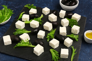 Wall Mural - Board with tasty cut feta cheese, salad leaves and black olives on blue grunge background