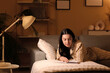 © Pixel-Shot - Young woman in pajamas reading book and lying on cozy sofa in living room at night