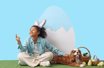 Canvas Print - Teen African-American girl with cute cavalier King Charles spaniel, Easter decor and bunny ears sitting on grass against blue background