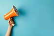 © Tasnim - Hand holding an orange megaphone against a blue background, symbolizing communication and announcement.