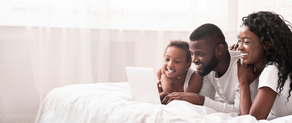 Wall Mural - Joyful black family of three using laptop in bed together, browsing internet or watching movie