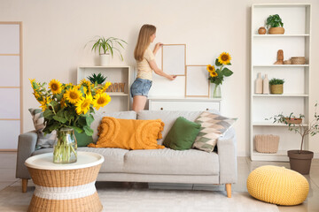 Wall Mural - Beautiful young happy woman with bouquets of sunflowers hanging blank frame at home