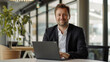© Tida - A businessman in a suit is sitting at a desk in a modern office, working on his laptop. He is smiling and looking directly at the camera, conveying confidence and professionalism. The image symbolizes