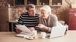 © Prostock-studio - An older couple sits at a kitchen table, reviewing documents while enjoying a cup of coffee. The man is holding a paper in his hands while the woman points to something on the paper.