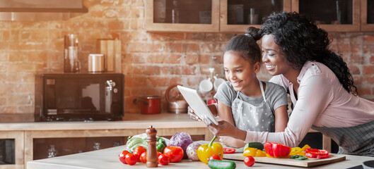 Wall Mural - Happy parenthood. Black woman and her cute daughter reading recipe on tablet, cooking at kitchen, copy space