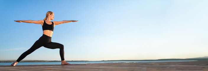 Beautiful Middle Aged Woman Practicing Yoga Outdoors, Standing In Warrior Pose, Portrait Of Fit Lady In Activewear Training On Wooden Pier Near Lake, Enjoying Healthy Lifestyle, Panorama, Copy Space