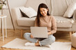 © Prostock-studio - A woman sits on a rug in a living room, using a laptop computer. She is smiling and appears to be focused on her work.
