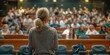 © Iryna - A woman stands in front of a large audience, giving a presentation. The audience is attentive and engaged, with many people looking at the woman. The atmosphere is serious and focused