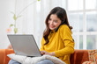 © 220 Selfmade studio - Asian young woman wearing a yellow sweater is multitasking by talking on her phone and using her laptop while sitting on a couch at home. She appears engaged and happy, working comfortably in a cozy