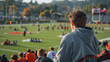 © khonkangrua - A teenager in a hoodie watches a high school football game from the stands, observing the field and players.