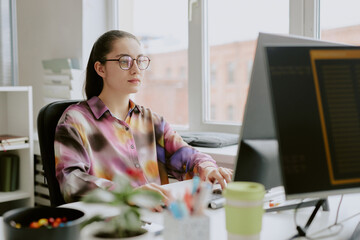 Young Caucasian female programmer dressed in spotted blouse sitting at work desk while typing on keyboard and looking directly at computer screen