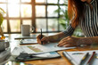 © sornram - Businesswoman analyzing financial chart at desk with laptop, pen, and papers in sunlit office.