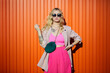 © wertinio - Elegant woman is wearing fashion pink or fuchsia color jacket, trousers on orange background. Fashion portrait of trendy woman wearing clothes, dark sunglasses, holding green bag.
