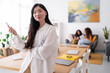 © Studio Marmellata - A woman in an orange shirt is using her smartphone while sitting on a desk in a bright office. She is smiling, and in the background, two other women are chatting on a couch.