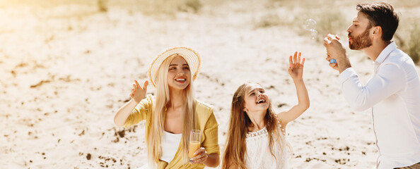 Wall Mural - Family picnic. Parents with daughter blowing soap bubbles, having rest in nature