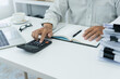 © PhotoJuthamat - office employee works on desk, using calculator to manage financial documents and accounts, preparing an income tax report in a busy office setting.