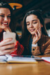 © BullRun - Positive female friends searching information for education using smartphone device and wifi connection in cafeteria, successful students enjoying break of studying watching funny video on telephone