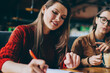 © BullRun - Positive carefree hipster girl writing essay with excited idea in textbook sitting near female friend with caffeine beverage, two happy successful women studying together at university cafeteria