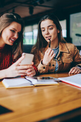 Wall Mural - Successful female students making research for examination using modern smartphone device and literature indoors, positive women checking updates for profile in social networks in break of studying
