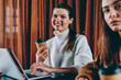 © BullRun - Portrait of positive smiling female teenager holding cup of takeaway coffee spending time with friend on leisure, two successful it professional enjoying distance job with netbooks at cafeteria