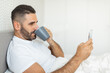 © Prostock-studio - A man with a beard sits in bed, drinking from a gray mug and using his phone, with white bedding and a white wall behind him.