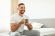 © Prostock-studio - A man sits on a bed, wearing a white t-shirt and grey sweatpants, holding a dark mug in his hands, smiling as he looks off to the side. The setting is a bedroom with a white bed, copy space
