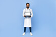 © Prostock-studio - Indian doctor with a beard, wearing a white coat, holds a laptop with blank screen in front of him and smiles at the camera.