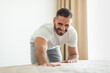 © Prostock-studio - A man in a white shirt and gray pants is bending over and making a bed. He is smiling and looks happy as he smooths the bedspread. The room is bright and airy, with a large window in the background.