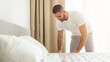 © Prostock-studio - A man in a white t-shirt and gray pants bends over to make his bed in a bright, modern bedroom. The bed is made with white sheets and pillows, and the room is decorated with neutral colors