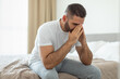© Prostock-studio - A man wearing a white t-shirt and gray pants is sitting on the edge of a bed. He has his hands on his face and is looking down, appearing to be in deep thought.