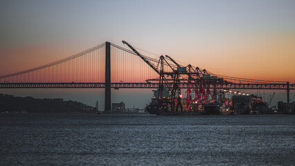 A panoramic view of a shipping port with cranes and a cargo ship against the backdrop of a bridge during sunset. The sky with vibrant colors creating a stunning silhouette of the industrial structures