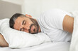 © Prostock-studio - A man with a beard is lying on his side in a bed, sleeping soundly. His eyes are closed, and his face has a relaxed expression. He is wearing a white t-shirt and is covered with a white blanket.