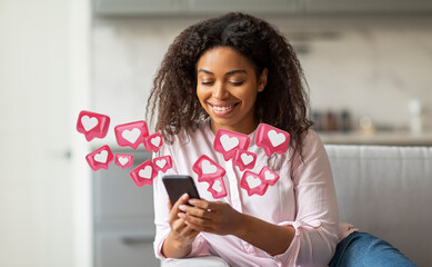 A young black woman with curly hair smiles as she uses her smartphone in her living room. Pink heart-shaped icons float around her, indicating social media likes and engagement.