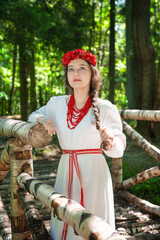 Wall Mural - Young calm Caucasian woman in a white Slavic dress stands near a birch bridge in a sunny forest