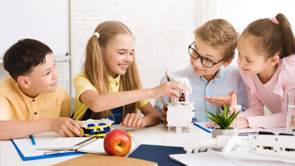 Poster - Four children, two boys and two girls, are working together on a robotics project in a classroom setting. They are all smiling and appear to be engaged in the task at hand