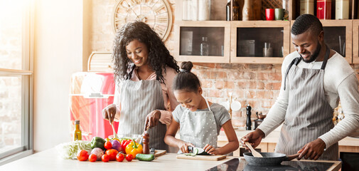 Wall Mural - Cute black little girl helping parents with dinner, spending time together at kitchen, empty space