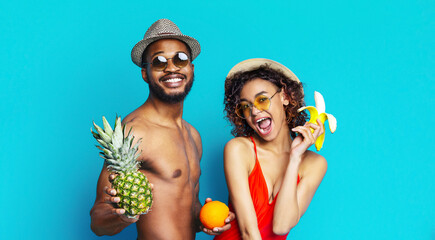 Wall Mural - Black man and woman are smiling and posing for the camera against a blue background. The man is wearing sunglasses and a hat and holding a pineapple. The woman is holding a banana and an orange.