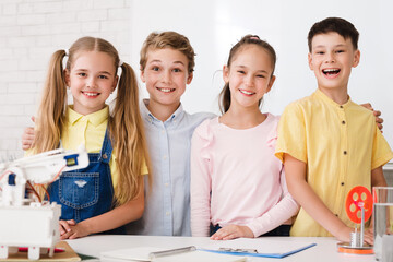 Aufkleber - Four young students, two boys and two girls, are smiling and working on a science experiment in a classroom. They are all wearing casual clothing and are standing around a table with a robot