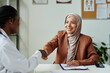 © pressmaster - Happy young Muslim female patient in hijab and casual attire shaking hand of African American clinician after signing medical agreement