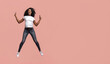 © Prostock-studio - This image shows a young African American woman with curly hair jumping in front of a plain pink background. She is wearing a white t-shirt, dark blue jeans, and white sneakers.