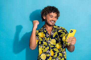 Sticker - Photo of satisfied man with wavy hairstyle dressed print shirt look at smartphone win lottery fist up isolated on blue color background