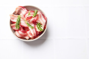 Wall Mural - Slices of raw bacon and rosemary in bowl on white tiled table, top view. Space for text