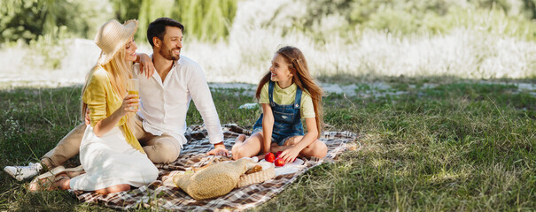 Wall Mural - Young family picnicking with their cute daughter, sitting on blanket in park