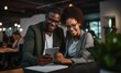 © Myimages - Successful two african american young businesspeople sitting on desk using digital tablet while colleague in background at office